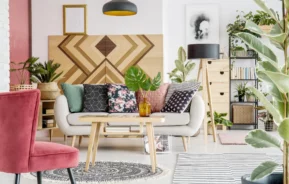 Foto de uma sala com decoração boho chic. A sala possui um sofá branco com almofadas coloridas e estampadas, uma poltrona rosa, plantas e um quadro com desenho étnico.