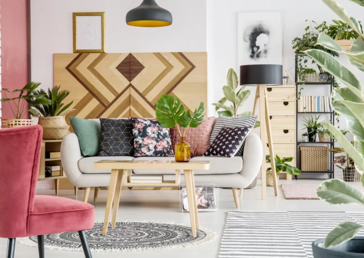 Foto de uma sala com decoração boho chic. A sala possui um sofá branco com almofadas coloridas e estampadas, uma poltrona rosa, plantas e um quadro com desenho étnico.