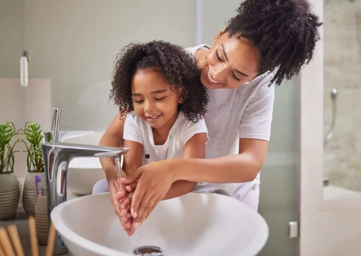 Imagem de uma mãe ajudando a filha a lavar as mãos em uma pia de banheiro para ilustrar matéria sobre a troca de titularidade Sabesp