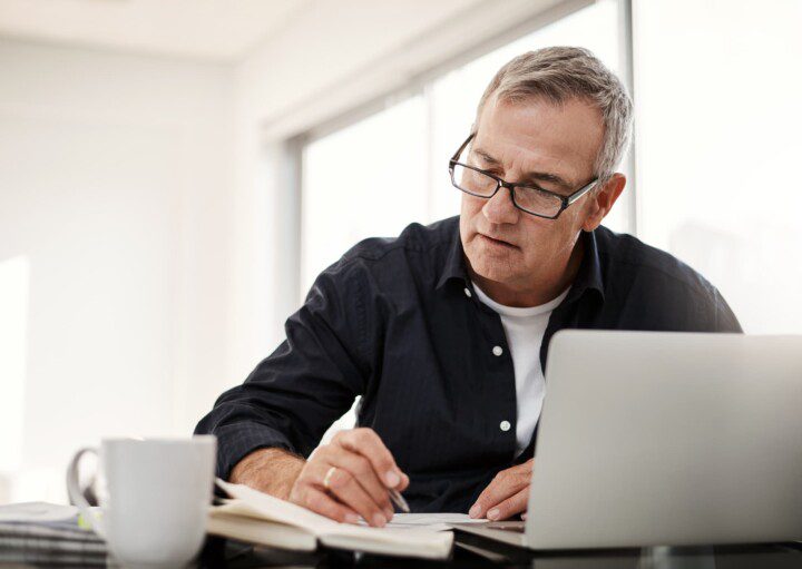 Foto que ilustra matéria sobre imposto sobre venda de imóvel mostra homem sentado à mesa com uma xícara de café, um notebook e fazendo anotações em um papel.