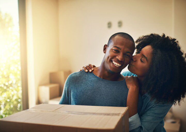 Foto que ilustra matéria sobre proposta de locação mostra um casal jovem que acaba de se mudar para uma casa nova. Na imagem, o homem sorridente ganha um beijo de sua parceira na bochecha.