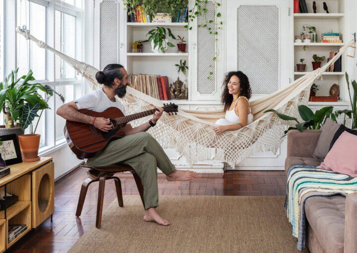 Foto mostra o casal Felippe e Carol na sala de seu apartamento no Paraíso, em São Paulo. Ele está sentado em um banco tocando violão, enquanto ela sorri sentada em uma rede de balanço