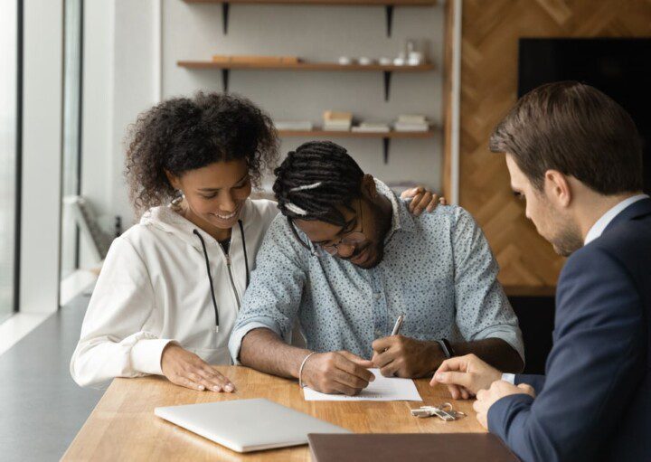 Foto que ilustra matéria sobre taxa de financiamento imobiliário mostra um casal de jovens assinando um contrato diante de um homem de terno