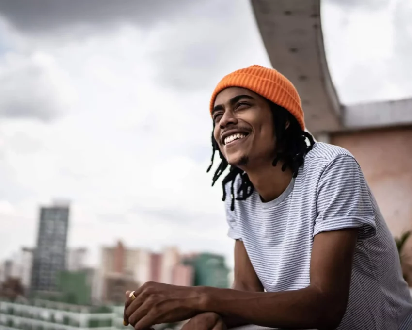 Imagem de um jovem encostado na sacada de um apartamento sorridente enquanto contempla a paisagem da varanda para ilustrar matéria sobre os melhores bairros para morar em SP