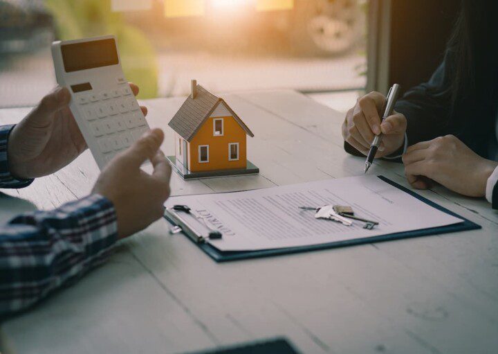 Pessoas analisam um contrato imobiliário com o auxílio de uma calculadora. Há, ao lado, a miniatura de uma casa.