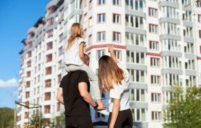 Em foto que ilustra matéria sobre como comprar um apartamento, um casal de costas aponta para um prédio ao fundo. A mulher é loira e o homem tem uma menina sentada em seus ombros.