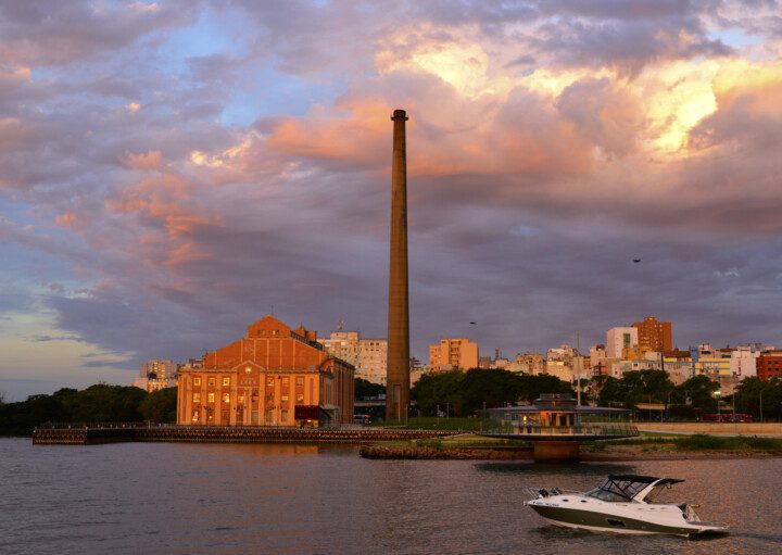 Foto que ilustra matéria sobre os melhores bairros de Porto Alegre mostra a Usina do Gasômetro, um dos pontos turísticos da cidade, às margens do Rio Guaíba, ao entardecer. Além do prédio e da chaminé ao lado, uma lança passa à frente.