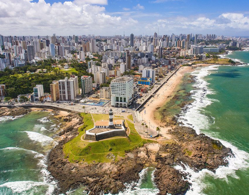 Foto que ilustra matéria sobre os melhores bairros de Salvador mostra os prédios do bairro da Barra vistos do alto, com o Farol da Barra e pedaços da praia em primeiro plano.