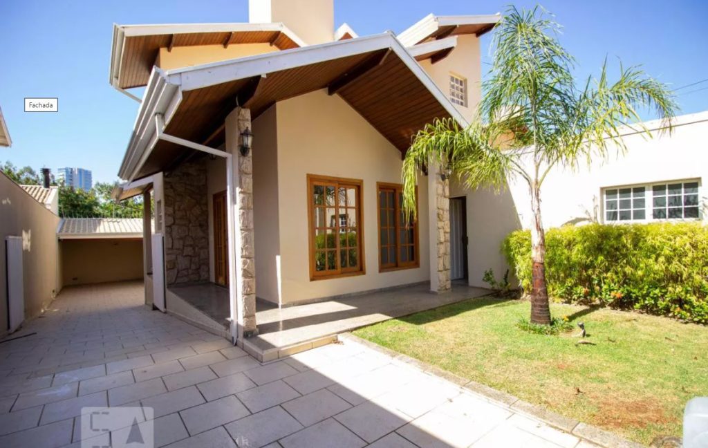 Uma casa clássica e planejada em um condomínio em Jundiaí. Há uma área externa gramada com uma palmeira e, aos fundos, a casa em tons amarelados. O sol ilumina a grama e parte da entrada. 