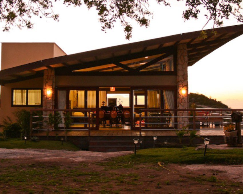 Foto que ilustra matéria sobre casas rústicas mostra uma casa no alto de uma estrutura rochosa, ao entardecer. A construção é cercada por uma varanda, tem um telhado em diagonal e uma bela vista para o pôr do sol.