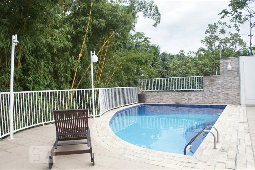 Piscina em um quintal com chão de pedras. Espreguiçadeira à frente.