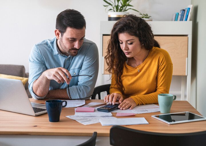 Foto que ilustra matéria sobre alugar ou financiar mostra um casal sentado a uma mesa, com um laptop e uma calculadora fazendo contas.