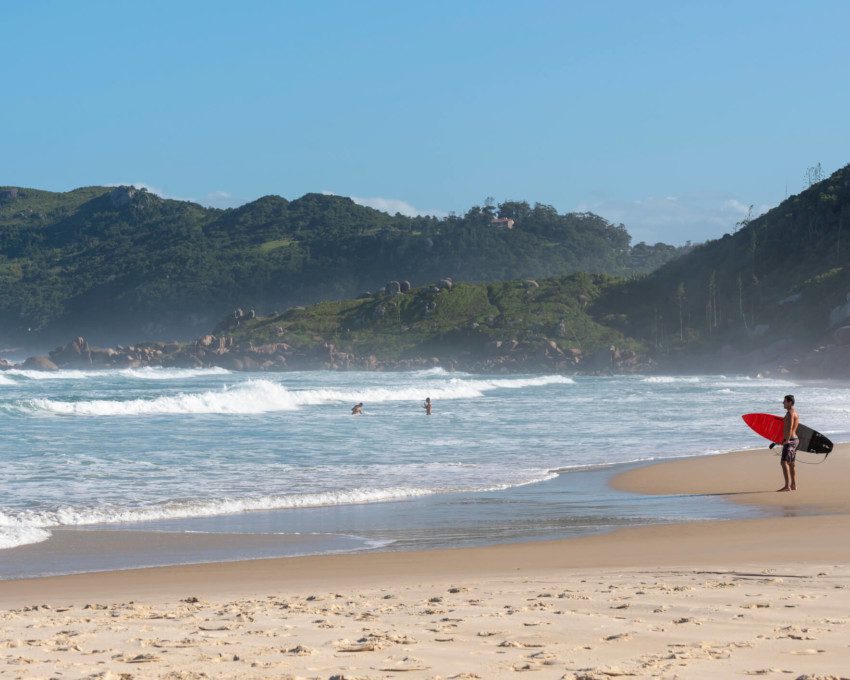 Foto que ilustra matéria sobre as praias de Florianópolis mostra um surfista de pé na areia, à direita da tela, com uma prancha de surfe debaixo do braço olhando para o mar na Praia da Galheta