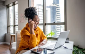 Imagem de uma mulher negra vestindo casaco laranja sentada na mesa da sala de casa olhando para a tela do computador sorrindo e atendimento ao telefone celular para ilustrar matéria sobre o seguro fiança locatícia