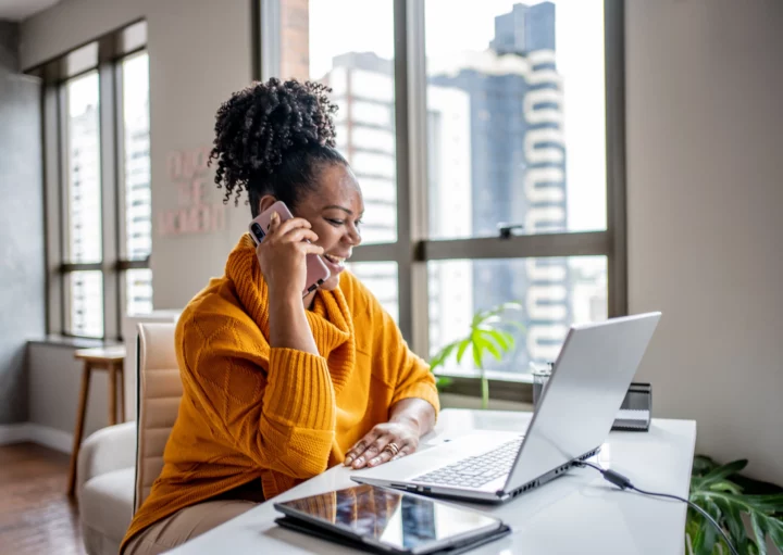 Imagem de uma mulher negra vestindo casaco laranja sentada na mesa da sala de casa olhando para a tela do computador sorrindo e atendimento ao telefone celular para ilustrar matéria sobre o seguro fiança locatícia