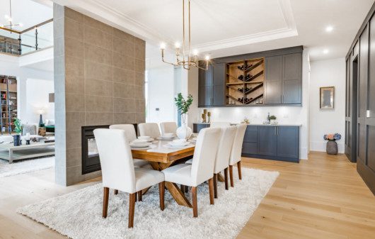 Foto que ilustra matéria sobre lustre para sala de jantar mostra uma mesa de jantar posta, em uma ampla sala, sobre um tapete felpudo branco e com oito cadeiras de encostos e assentos brancos a sua volta.