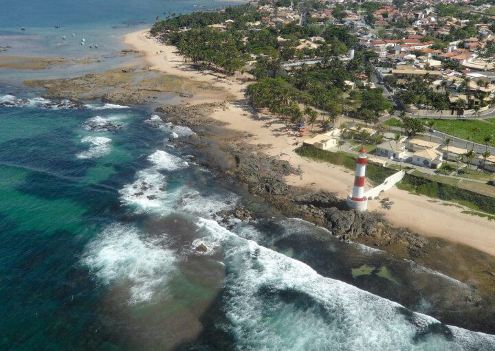 Foto que ilustra matéria sobre as praias de Salvador mostra uma vista aérea da Praia de Itapuã, com o mar ocupando parte da tela do meio para a esquerda, o famoso farol de Itapuã no meio e a faixa de terra mais para o canto superior direito da imagem.