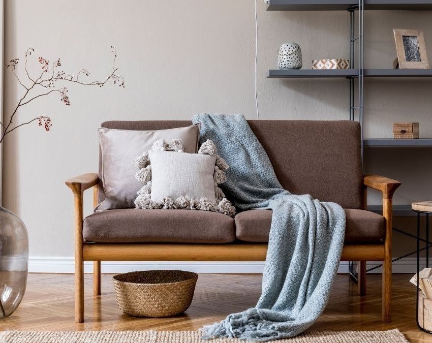 sala de estar decorada no estilo japandi. Sofá de dois lugares, cinza, com manta azul caida no chão. Ao lado, vaso com flores e mesinha redonda. Atrás, prateleira com objetos decorativos