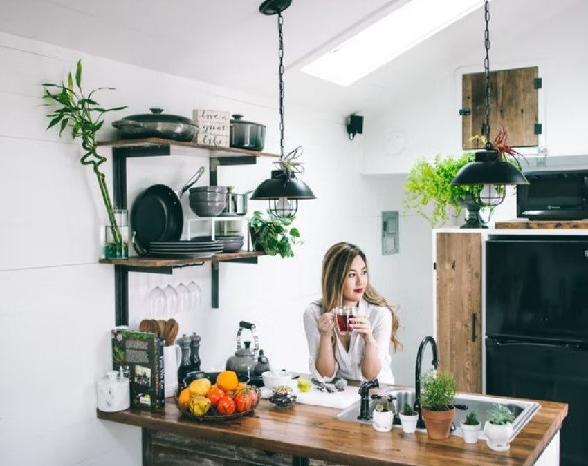 A foto mostra uma mulher sentada à mesa de uma cozinha pequena e bem decorada. Na imagem há uma bancada com pia, frutas, plantas, livro, chaleira e temperos. Há também na imagem uma prateleira com utensílios de cozinha (pratos, panelas e outros), além de uma geladeira.