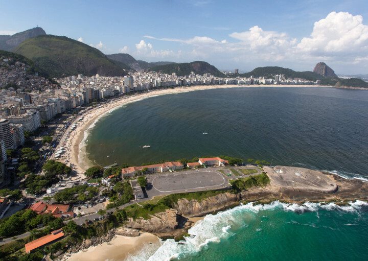 Foto que ilustra matéria sobre as praias do Rio de Janeiro mostra uma visão aérea de toda a orla da praia de Copacabana, a começar pelo Forte de Copacabana, em primeiro plano, mais abaixo na tela, seguido de toda a faixa de areia e boa parte do mar, com os prédios à esquerda e montanhas ao fundo. Entre elas os morros do Corcovado e do Pão de Açúcar