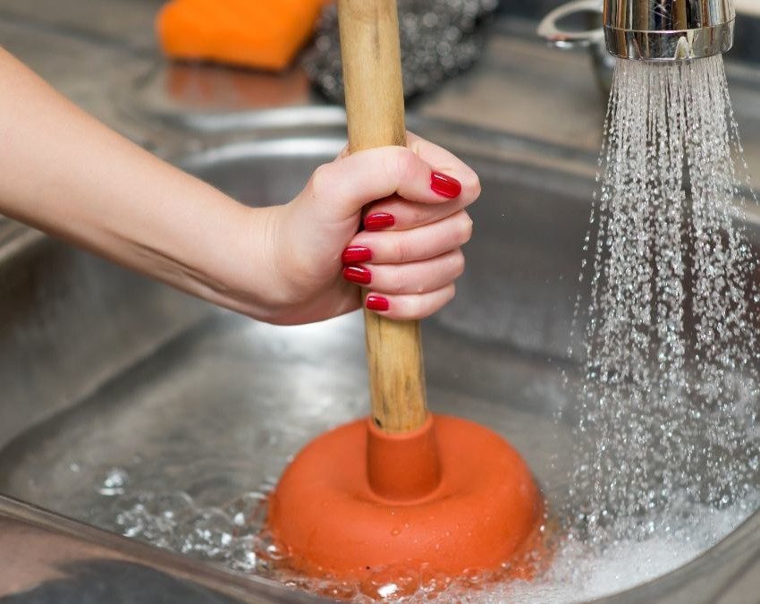 Mão com unhas pintadas de esmalte segurando um desentupidor dentro de uma pia de cozinha entupida, ensinando como desentupir pia de cozinha