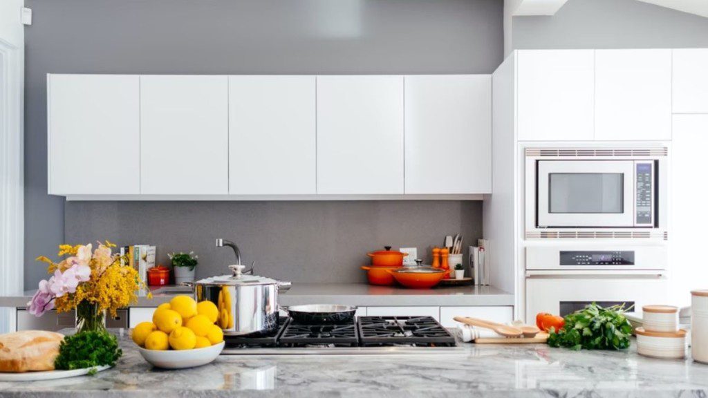 A foto mostra uma cozinha com frutas, temperos e panelas decorando o ambiente. Os itens estão sobre a bancada e também sobre a pia. Há também armário, forno, fogão e utensílios como colheres e panelas. 