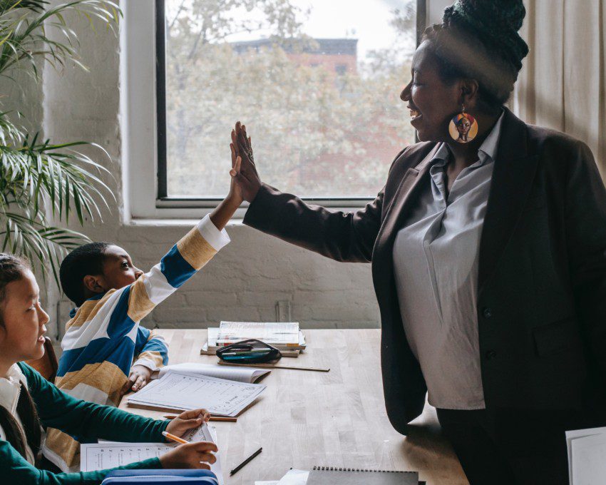 Foto que ilustra matéria sobre melhores escolas em Recife mostra uma professora negra fazendo um gesto de toca aí com um aluno, enquanto uma aluna ao lado observa