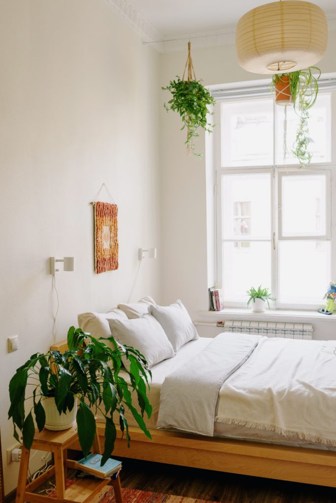 Imagem de um quarto de casal com uma cama de madeira e o jogo de cama todo branco. Ainda tem duas luminárias brancas em cada lado da cama, um banco servindo de mesinha de cabeceira e algumas plantas penduradas no teto.