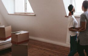 Foto que ilustra matéria sobre o Censo QuintoAndar com foco em desejos de mudança de casa mostra um casal abraçado em um quarto, de costas, olhando por uma janela. E ao lado esquerdo uma cama com algumas caixas de papelão em cima em no chão à frente dela