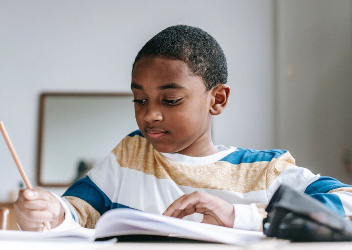 Foto que ilustra matéria sobre escolas em Santo André mostra um menino negro sentado à uma mesa enquanto escreve de lápis em um caderno.