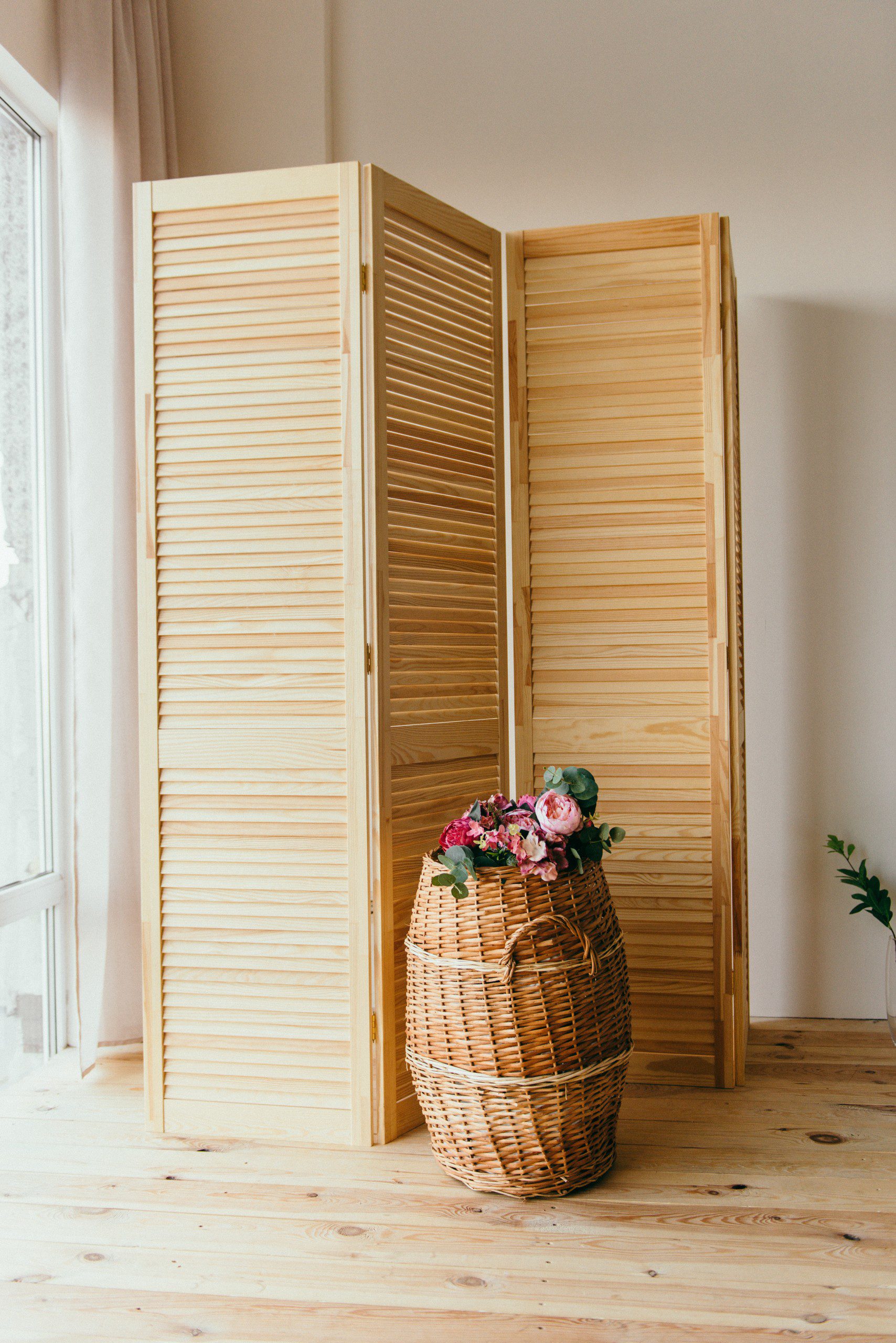 Imagem de um biombo de madeira ao lado de um cesto também de madeira em um ambiente com parede bege. 