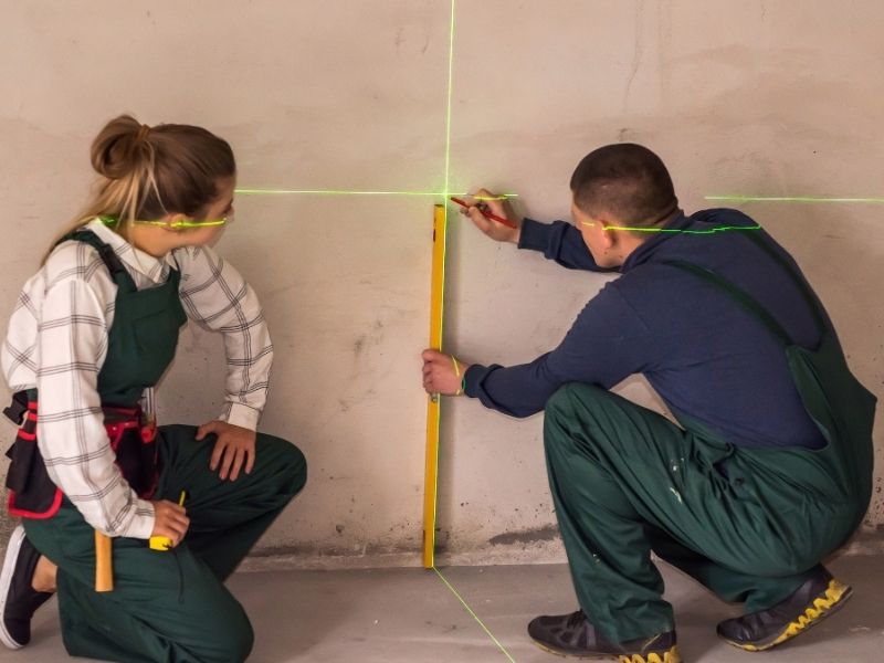 Foto que ilustra matéria sobre como calcular metro quadrado mostra homem e mulher medindo um ambiente, com luzes laser iluminando os quadrantes