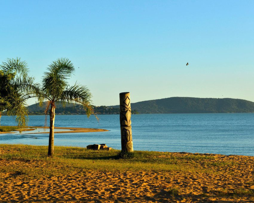 Foto que ilustra matéria sobre as praias de Porto Alegre mostra um trecho da praia de Ipanema, onde aparecem uma faixa de areia em primeiro plano, alguns coqueiros à esquerda, o espelho d’água no meio da imagem e pequenos morros ao fundo.