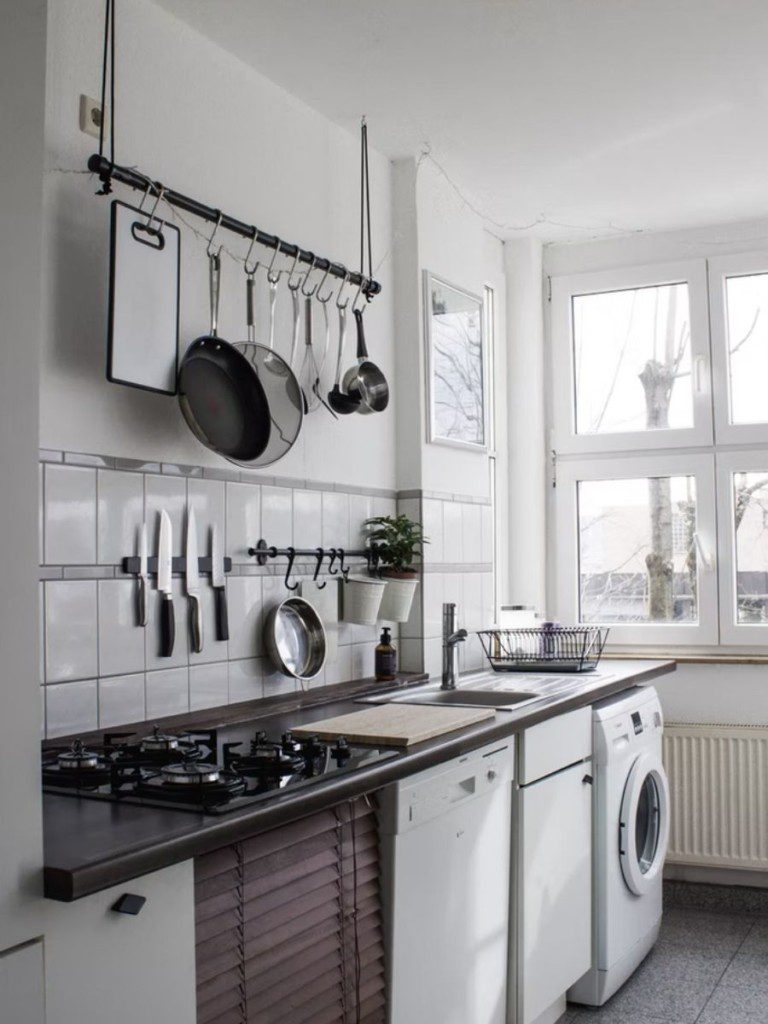 A imagem mostra uma cozinha com área de serviço integrada. Nela há uma máquina de lavar, uma bancada com armário embutido, uma pia, um fogão e diferentes utensílios de cozinha em cima da bancada.