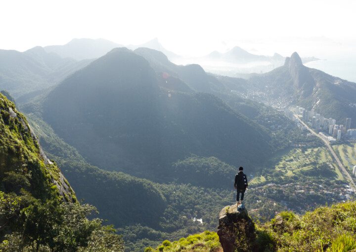 Imagem panorâmica de um homem de costas em cima da Pedra da Gávea olhando as montanhas do Rio de Janeiro para ilustrar matéria sobre parques no Rio de Janeiro