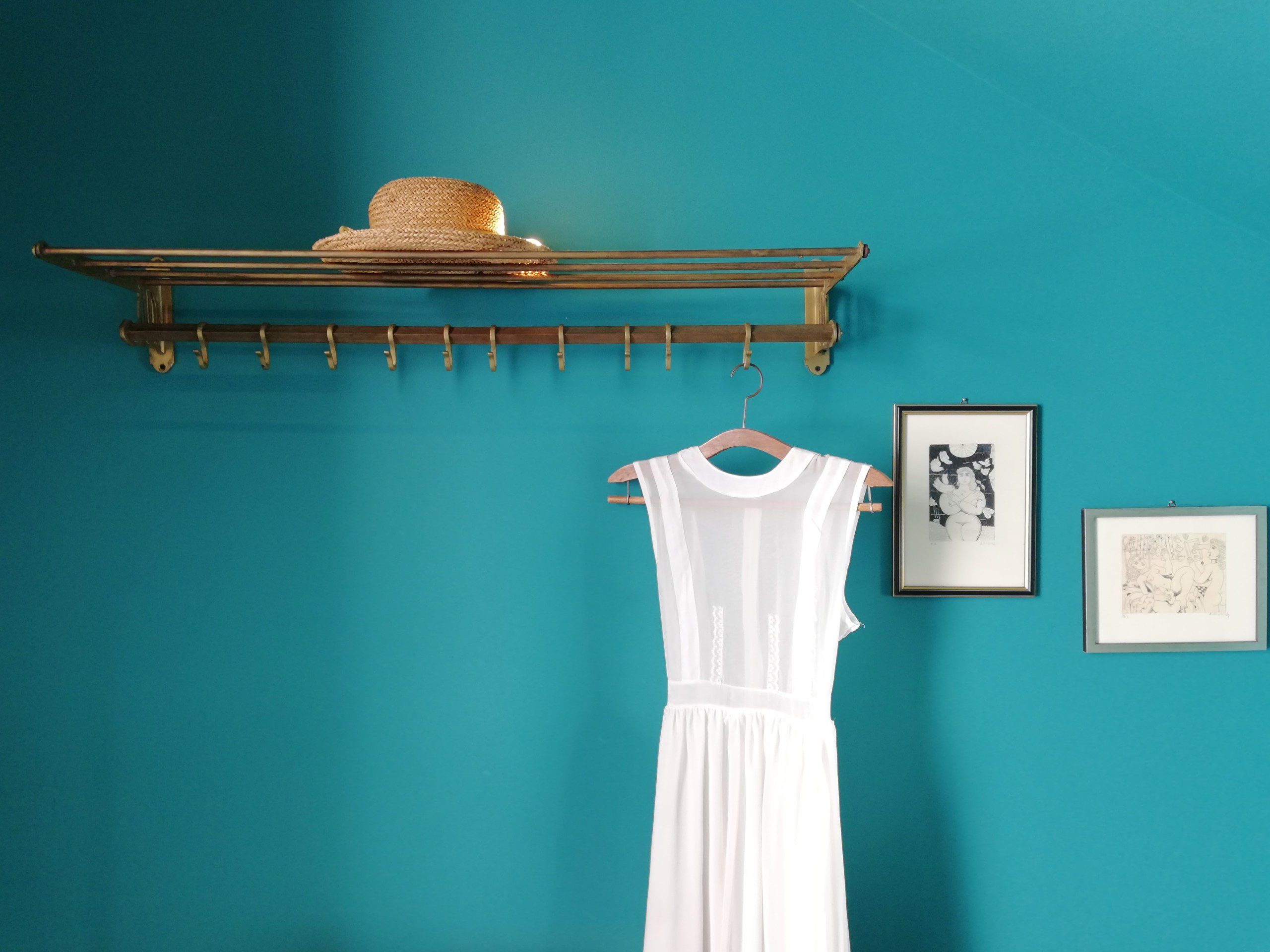 Imagem de uma parede azul turquesa com quadros decorativos, um chapéu em cima de uma prateleira com um vestido no cabide pendurado em um gancho.  
