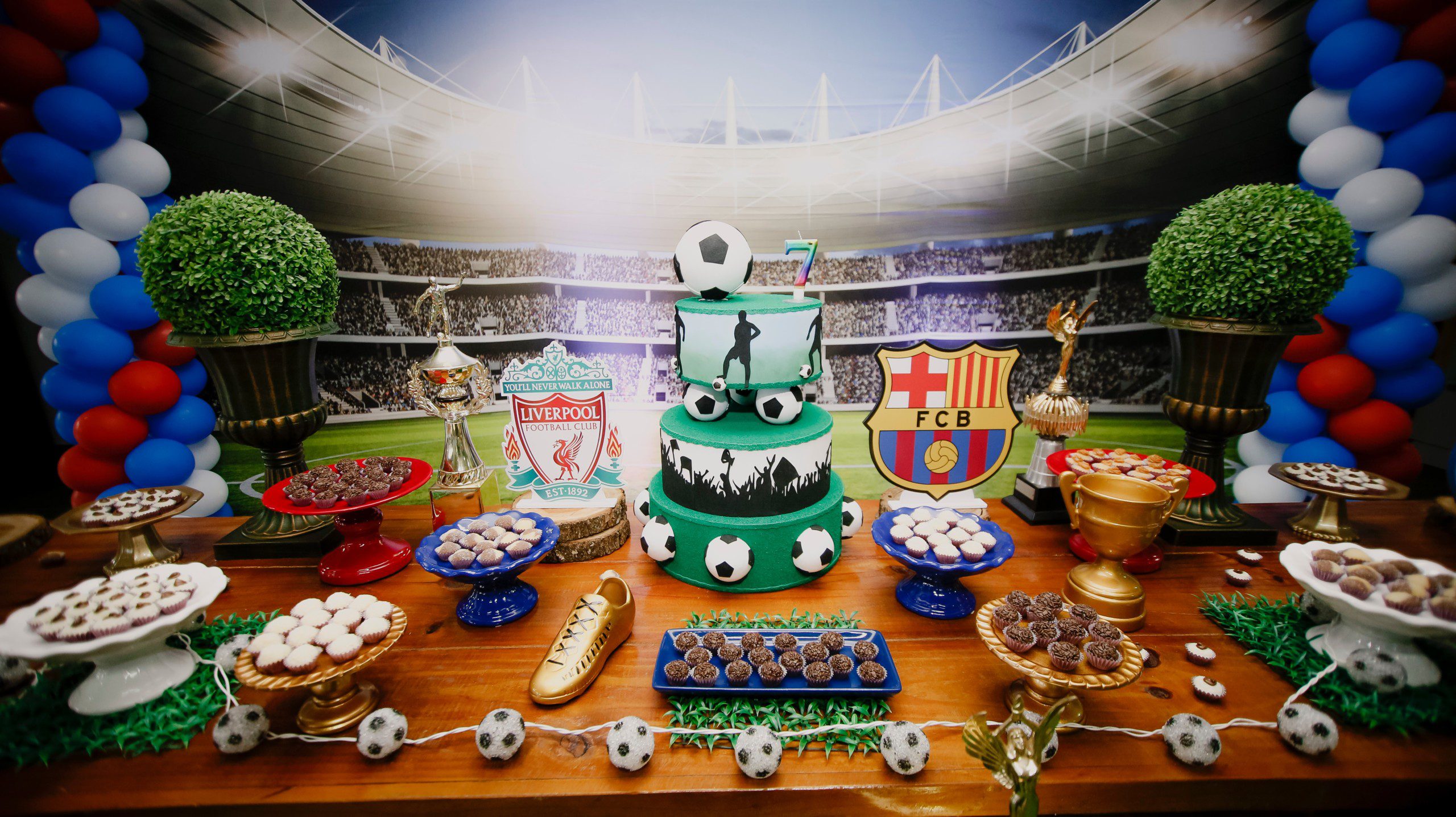 Imagem de uma decoração de festa simples com a temática de futebol com bolo de três andares, balões e diversos brigadeiros. 