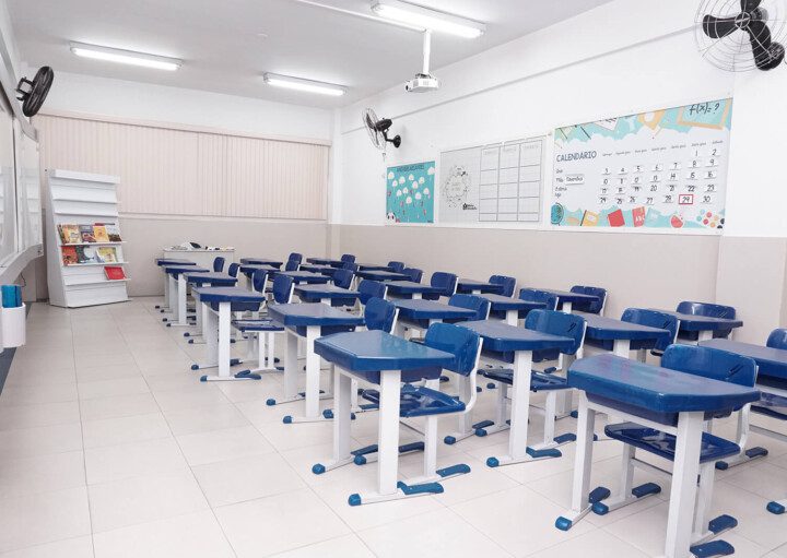 Foto que ilustra matéria sobre escolas particulares em Guarulhos mostra uma sala de aula da escola Mater Amabilis, em Guarulhos. A sala tem paredes brancas e chão claro. E as carteiras, conjuntos de cadeira e mesa, aparecem enfileiradas, lado a lado e uma em frente à outra. Elas têm estrutura branca de ferro e revestimento azul.