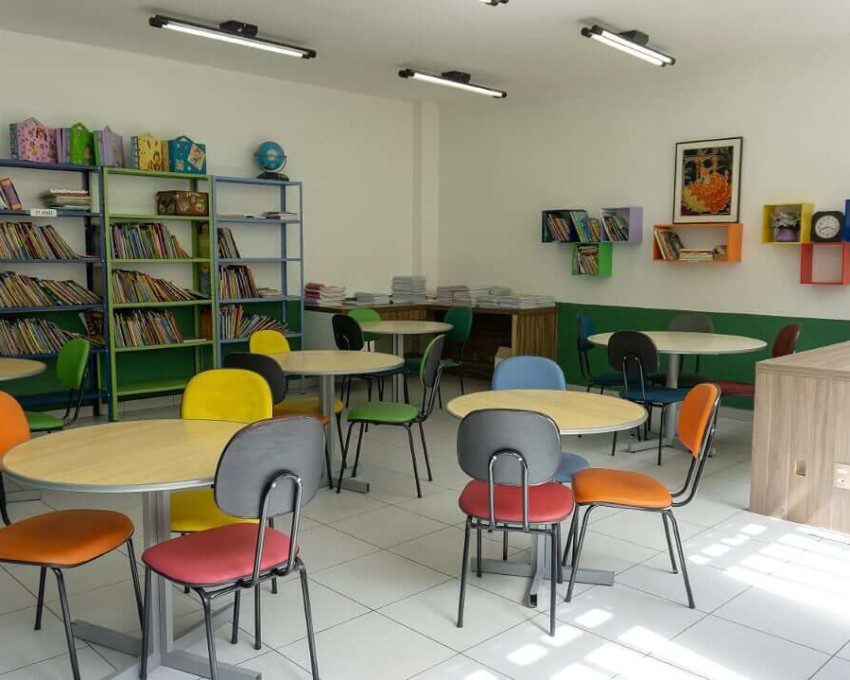 Foto que ilustra matéria sobre escolas em Osasco mostra uma sala de estudos do Colégio Cunha Carvalho, com diversas mesas redondas de madeira cercadas por cadeiras com estofados de diferentes cores. Ao fundo, há estantes com diversos livros.