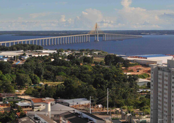 Foto que ilustra matéria sobre os melhores bairros de Manaus mostra ao fundo um grande espelho d’água formado pelo Rio Negro com uma ponte cruzando a image de uma ponta à outra. Do centro da foto para baixo, à margem do rio, há uma faixa de terra com árvores, algumas casas e um prédio.