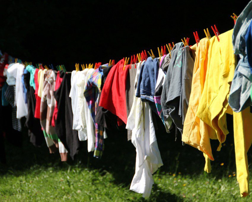 Foto que ilustra a matéria com dicas de como tirar mofo de roupa mostra um varal no sol com roupas de diferentes cores. As roupas estão penduradas com prendedores também em cores variadas. E o varal está em uma área externa da casa com grama.