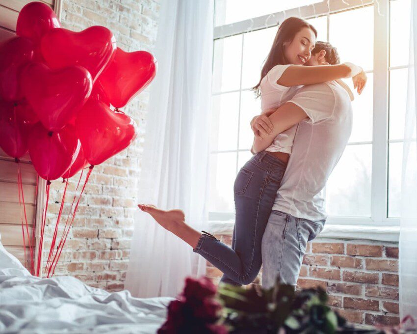 Casal de namorados se abraçando. O quarto está com decoração para o dia dos namorados.