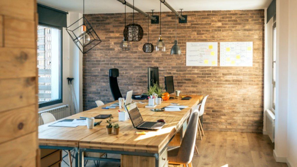 A imagem mostra uma sala de escritório em tons amadeirados com mesa ampla, cadeiras ao redor, iluminação pendente e parede de tijolos. Há também na imagem telefone e notebooks em cima da mesa.