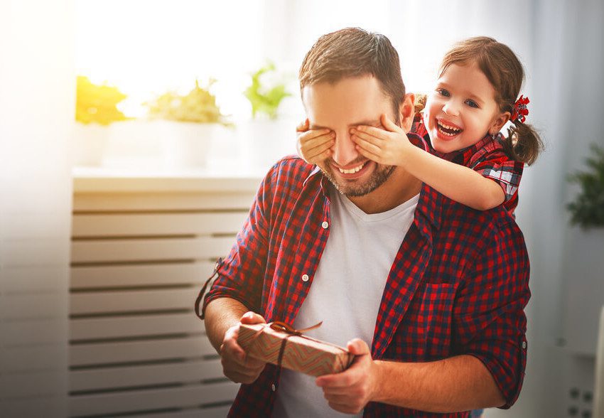 Foto que ilustra matéria sobre decoração para Dia dos Pais mostra um homem sorridente segurando um embrulho pequeno de presente nas mãos enquanto uma menina, também sorridente, está atrás e tampando os olhos dele com as mãos