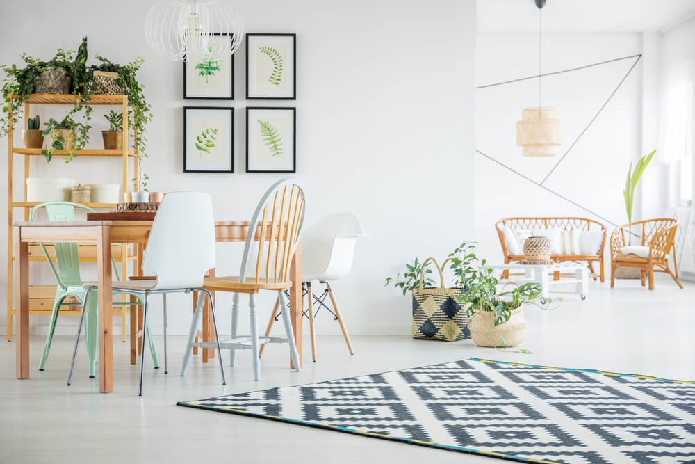 Na imagem tem uma mesa de jantar retangular com quatro cadeiras ao redor, uma estante de madeira, um conjunto de 4 quadros decorativos, tapete, sofá e poltrona de madeiras, além de diferentes plantas.