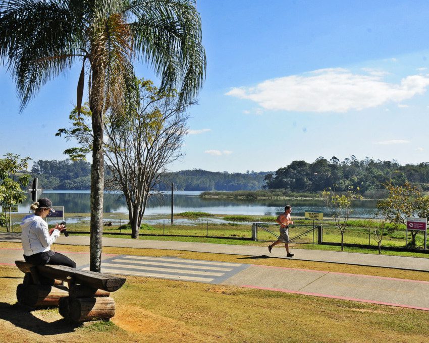 Foto que ilustra matéria sobre o que fazer em Jundiaí mostra uma paisagem do Parque da Cidade, com uma mulher sentada em um banco enquanto olha uma câmera fotográfica à esquerda, em primeiro plano, uma pista de corrida, com um homem se exercitando, e ao fundo um espelho d’água seguido de árvores em um dia de céu azul e poucas nuvens.