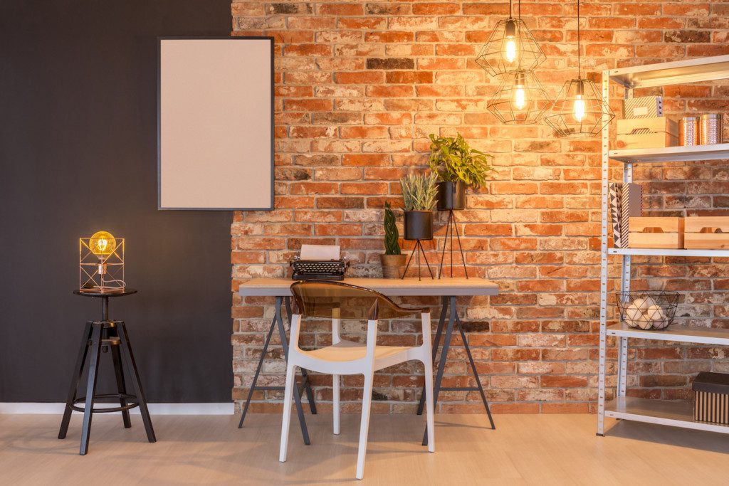 Na imagem há uma parede que é metade preta, metade com tijolos aparentes. Há também um lustre de chão, uma mesa de trabalho, cadeira, estante de ferro e um quadro em branco, além de outros elementos decorativos como plantas, caixas organizadoras e máquina de escrever.