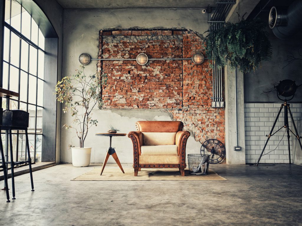 A imagem mostra uma parede com tijolos aparente, onde há um lustre com tubulação também aparente. Tem também uma poltrona com uma mesa ao lado, tapete, lustre de chão e uma planta decorativa.