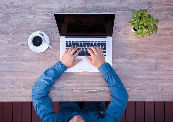 Imagem que ilustra matéria sobre comprar ou investir em fundos imobiliários mostra um homem sentado em frente a um computador com um café do lado esquerdo e um vaso de planta do lado direito.