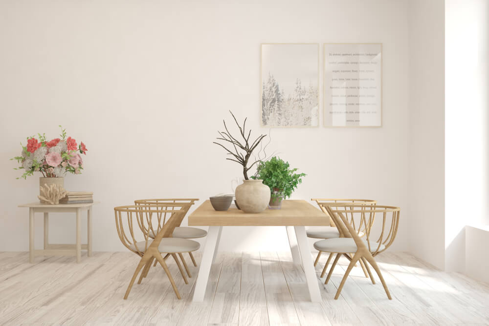 A imagem mostra uma sala de jantar onde há uma mesa retangular com quatro cadeiras ao redor. Há também uma mesa menor de apoio com flores em um vaso em cima dela e quadros decorativos na parede. 